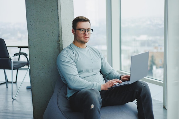 Um jovem gerente de óculos e roupas casuais senta-se em uma cadeira macia e trabalha em um laptop Espaço de trabalho para trabalho remoto