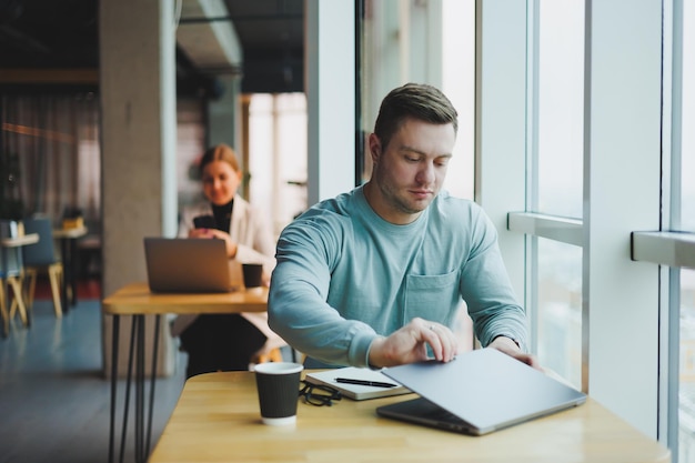 Um jovem gerente do sexo masculino senta-se em um escritório com grandes janelas e trabalha em um laptop Um belo empresário está trabalhando em sua lição de casa em um café