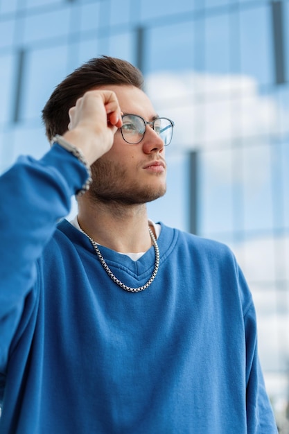 Um jovem hipster bonito e elegante em um elegante suéter azul usa óculos na cidade perto de um prédio comercial