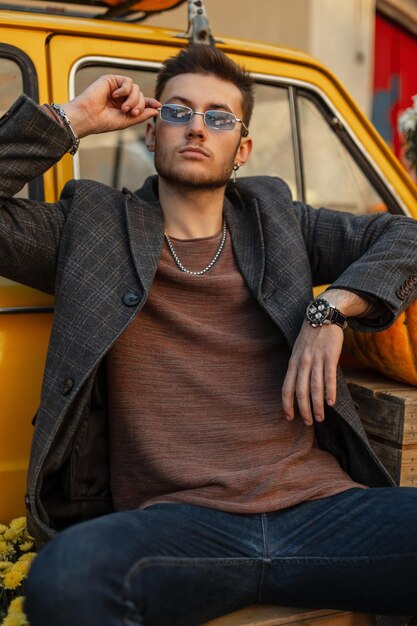 Foto um jovem hipster moderno e elegante com jeans e moletom da moda usa óculos escuros e descansa perto de um carro retrô amarelo