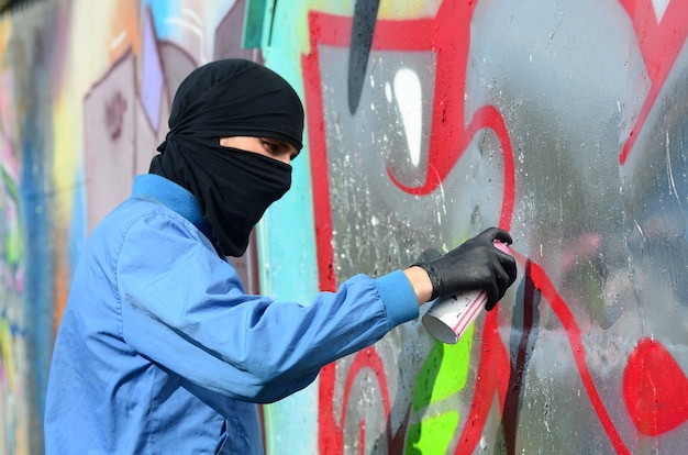 Um jovem hooligan com uma cara escondida pinta graffiti em uma parede de metal. Conceito de vandalismo ilegal