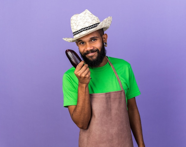 Um jovem jardineiro afro-americano satisfeito com um chapéu de jardinagem e segurando uma berinjela