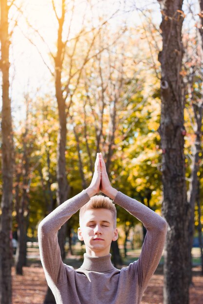 Um jovem loiro, vestido com um suéter bege, está com os olhos fechados, esticando os braços e dobrando-os