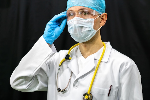 Um jovem médico em um fundo preto Um médico usando uma máscara protetora e luvas durante a pandemia de coronavírus