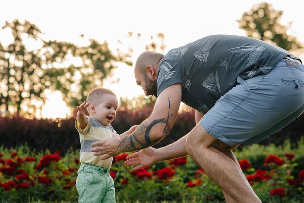 Um jovem pai barbudo ajuda e ensina seu filho a dar os primeiros passos durante o pôr do sol no parque na grama.