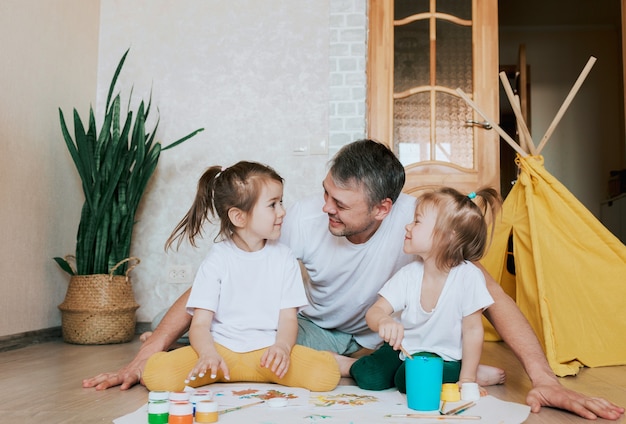 Um jovem pai carinhoso aprende a pintar com suas filhas, um homem deita-se no chão e ensina as meninas a desenhar flores e objetos de decoração.