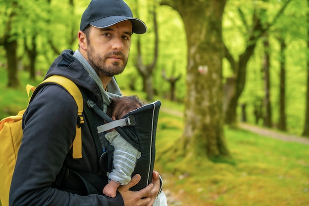 Um jovem pai com uma mochila amarela e um boné preto caminhando com o filho recém-nascido na mochila em um caminho na floresta