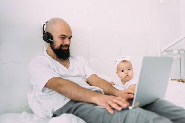 Um jovem pai está sentado em uma cama com uma criança juntos em casa em um computador Um pai trabalha em um laptop enquanto sua filha olha