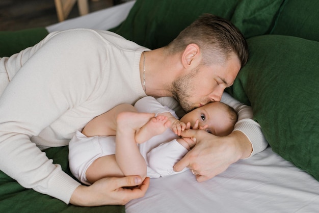 Um jovem pai feliz deita na cama com seu filho de seis meses na cama e o beija