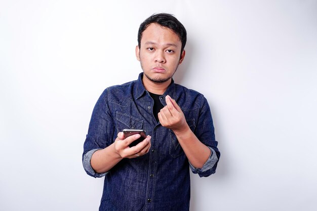 Um jovem pensativo vestido com camisa azul enquanto olha de lado segurando seu telefone isolado pelo fundo branco