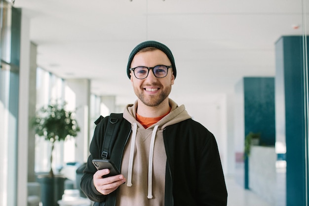 Um jovem positivo com um smartphone nas mãos está olhando para a câmera Um jovem de óculos e boné está parado em um escritório