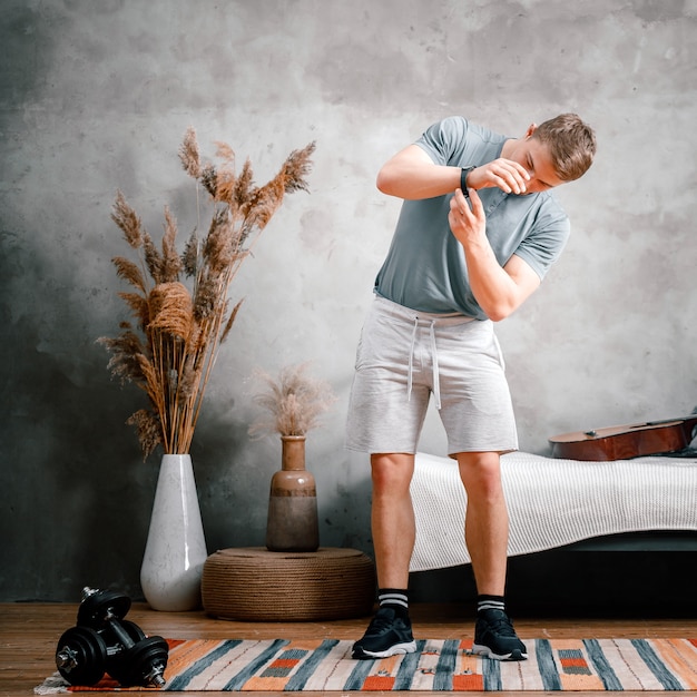 Um jovem pratica esportes em casa, treina online pelo telefone. A atleta coloca um relógio esportivo, se prepara para os treinos no quarto, ao fundo tem uma cama, um vaso, um tapete.