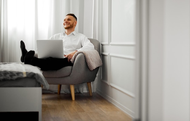Um jovem ri trabalhando em um quarto branco com um laptop