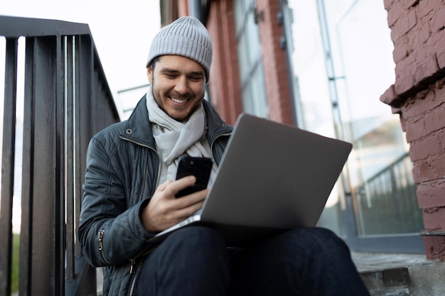 Um jovem senta-se na escada com um laptop e olha para o telefone com um sorriso