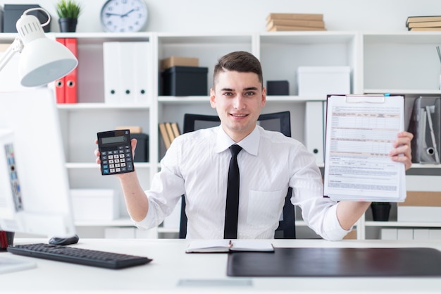 Um jovem sentado em uma mesa de computador no escritório e segurando um tablet e uma calculadora.