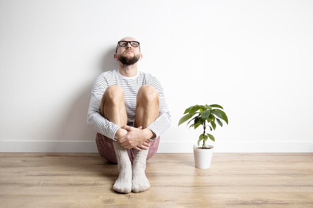 Um jovem sentado no chão de madeira olha para cima ao lado de uma flor em um vaso