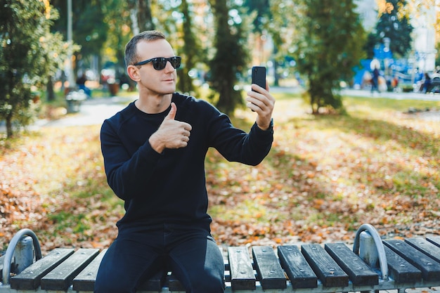 Um jovem solitário de suéter preto senta-se no parque em um banco e fala ao telefone Um dia quente de outono