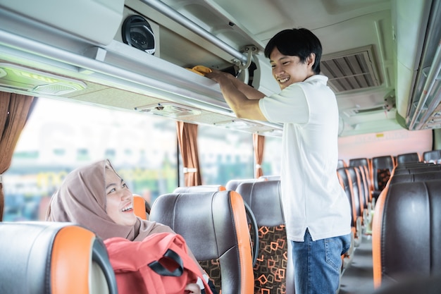 Um jovem sorri enquanto ajuda uma mulher com um lenço na cabeça a colocar sua bolsa em uma prateleira enquanto está de pé no ônibus