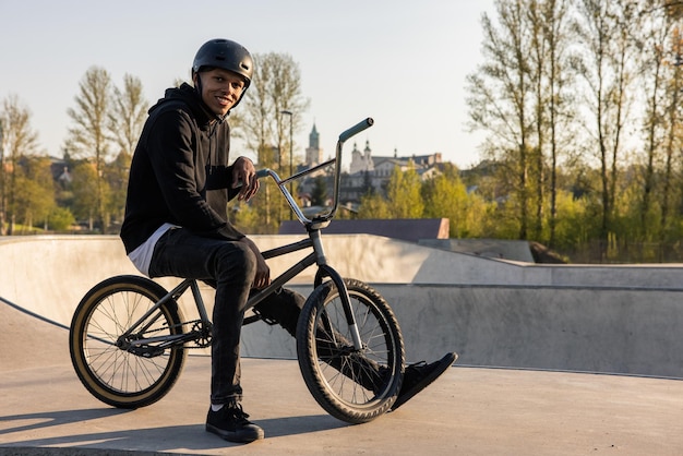 Um jovem sorridente está usando um capacete na cabeça e de pé em uma rampa com uma bicicleta para fazer truques