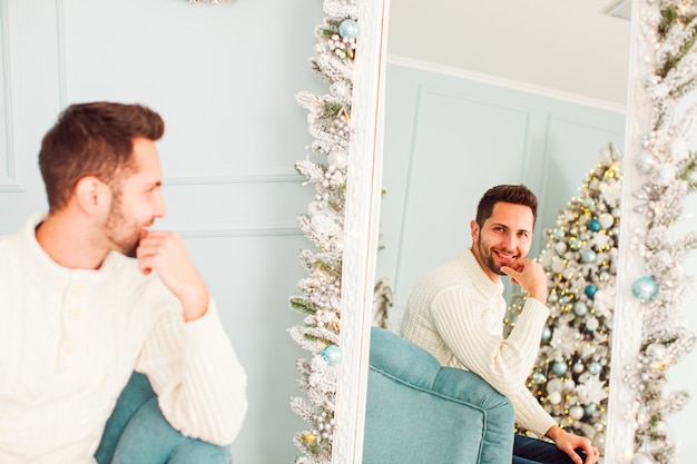 Um jovem sorridente, sentado na poltrona azul claro, olhando para si mesmo no grande espelho no estúdio decorado de Natal com abeto interior de férias e luzes de férias