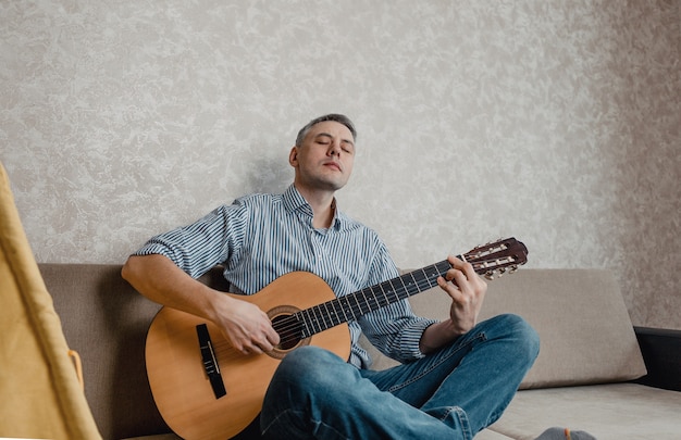 Um jovem toca violão sentado em um sofá em uma sala bem iluminada. Um instrumento musical para o conceito de lazer ou hobby. entretenimento caseiro