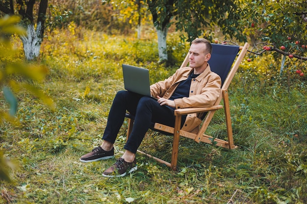 Um jovem trabalha em um laptop ao ar livre Um jovem freelancer descansa na floresta Trabalho remoto recreação ativa no verão Turismo pessoas conceito homem sentado em uma cadeira ao ar livre