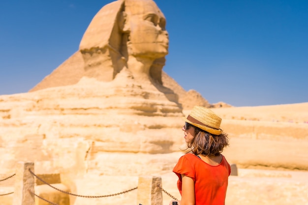 Um jovem turista na Grande Esfinge de Gizé vestido de vermelho e com um chapéu, de onde saem as miramides de Gizé. Cairo, Egito