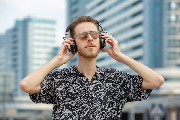 Foto um jovem usando fones de ouvido e óculos escuros ouve música no contexto de edifícios modernos