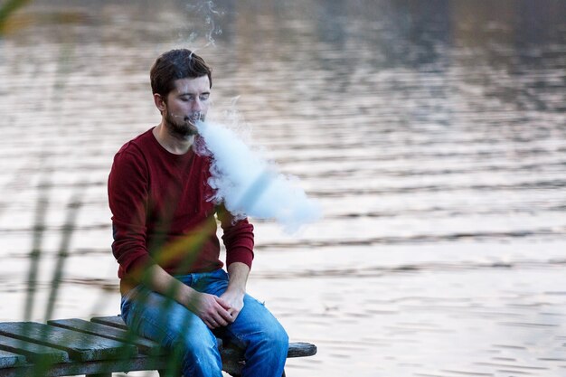 Um jovem vaper senta-se em uma ponte em um dia de outono e fuma