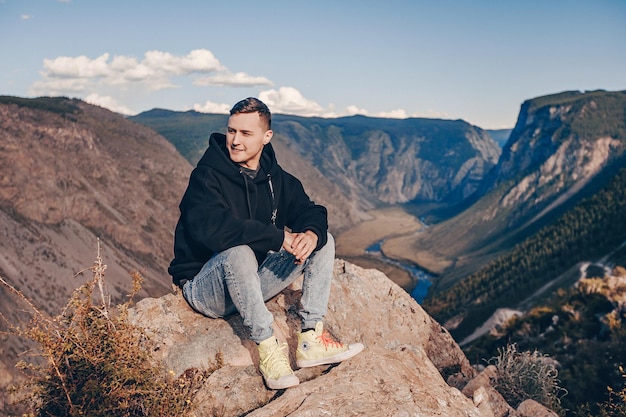 Um jovem vestido com um capuz preto senta-se em um paralelepípedo no penhasco de uma montanha e sorri olhando para longe. Atrás há uma paisagem natural de montanha com um riacho fluindo ao longe.