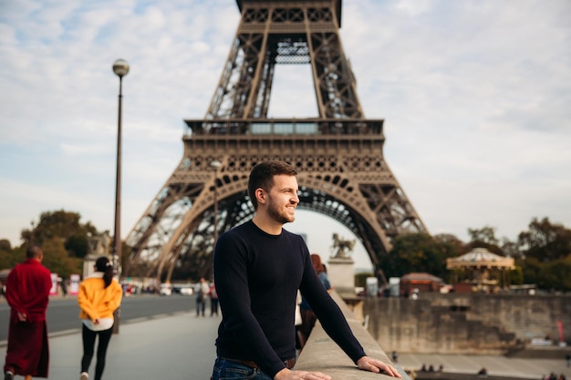 Um jovem vestindo uma jaqueta azul escura está de pé no fundo da torre eiffel ensolarada