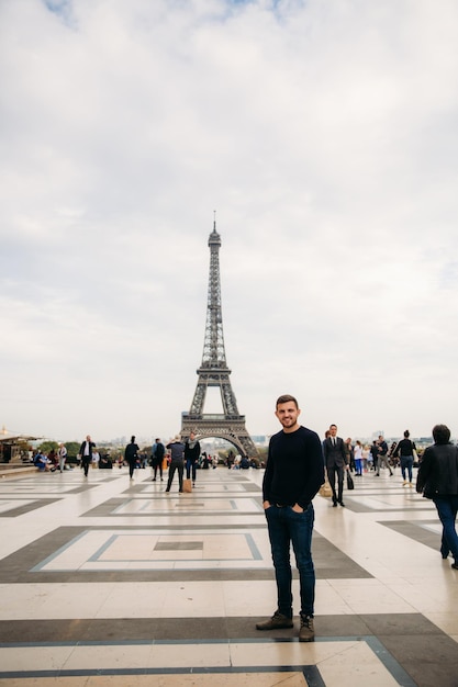 Um jovem vestindo uma jaqueta azul escura está de pé no fundo da Torre Eiffel O clima ensolarado é outono