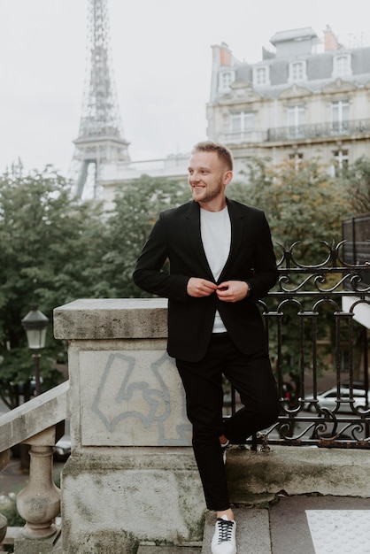 Um jovem vestindo uma jaqueta preta escura está de pé no fundo da Torre Eiffel