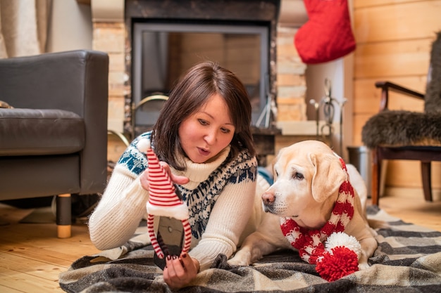 Um labrador retriever dourado em um lenço de Natal encontra-se com uma mulher asiática de meia-idade em um cobertor em frente a uma lareira em uma casa de campo.