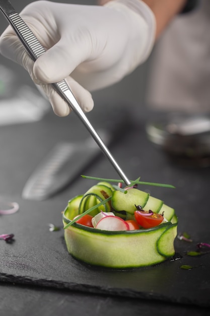 Um lanche de vegetais feito de vegetais frescos como pepino, tomate, queijo feta e microgreens. Um chef enluvado espalha cebolas verdes com uma pinça em um prato gourmet