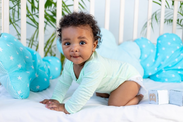 Um lindo bebê africano senta-se ou engatinha em uma cama em casa em uma cama de algodão azul e sorri.