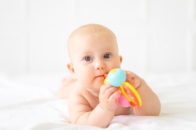 Um lindo bebê alegre e saudável em uma fralda está deitado em uma cama com roupa de cama branca, a criança está segurando um brinquedo olhando para a câmera