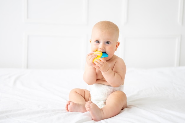 Foto um lindo bebê alegre e saudável em uma fralda está sentado em uma cama com roupa de cama branca, a criança está segurando um brinquedo olhando para a câmera