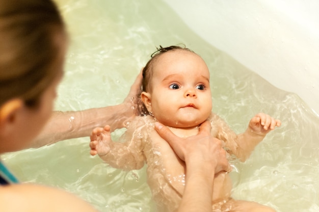 Foto um lindo bebê pequeno e fofo de olhos grandes tomando banho em água morna nas mãos de uma mãe carinhosa