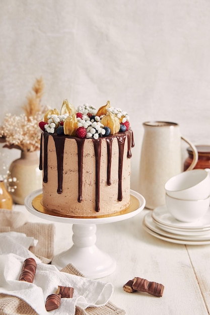 Um lindo bolo de chocolate com decorações de flores e bagas em um suporte de bolo branco em uma cozinha brilhante