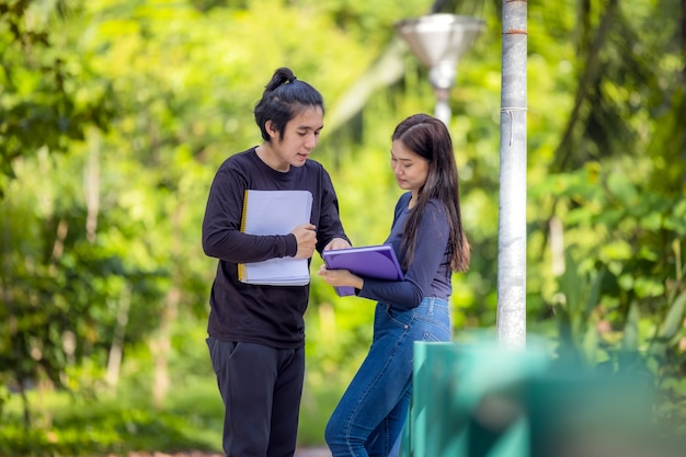 Um lindo casal de estudantes está parado no parque do campus de uma universidade com cadernos e livros nas mãos, fazendo uma pausa juntos.