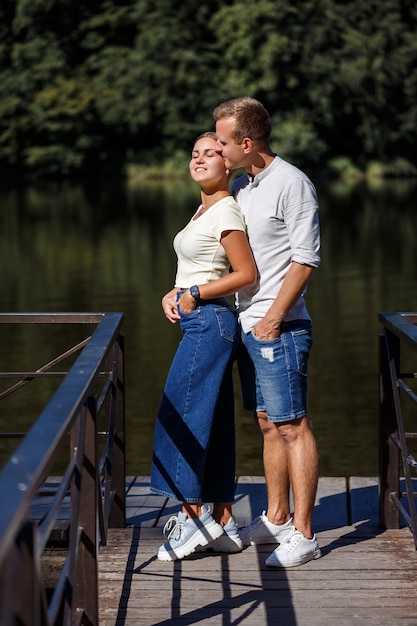 Um lindo casal jovem fica na margem do rio que eles abraçam. Casal feliz de férias à beira do lago