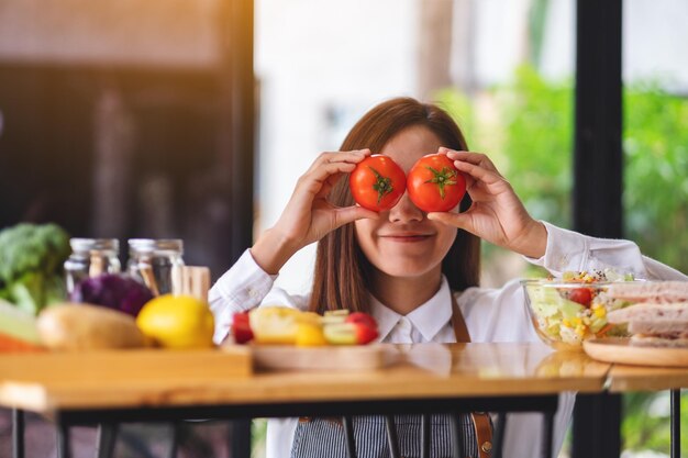 Um lindo chef feminino segurando e cobrindo os olhos com tomates enquanto cozinha na cozinha