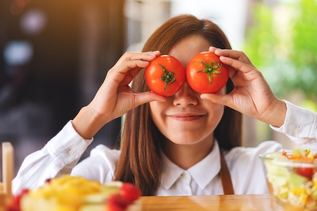 Um lindo chef feminino segurando e cobrindo os olhos com tomates enquanto cozinha na cozinha