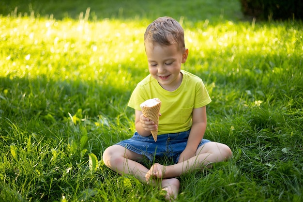 Um lindo garotinho com olhos azuis e cabelos loiros come sorvete no parque se senta na grama verde sorri se alegra