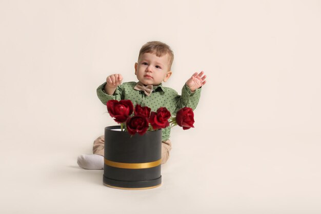 Um lindo garotinho em uma camisa verde sentado em um fundo branco ao lado de uma caixa com rosas vermelhas