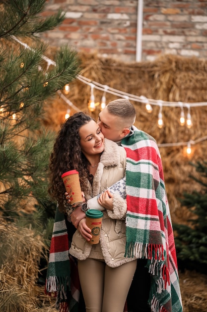 Um lindo jovem casal com roupas brancas está sentado perto de uma árvore de Natal no jardim. Homem e mulher felizes, romance, celebração de Natal, diversão, amor.
