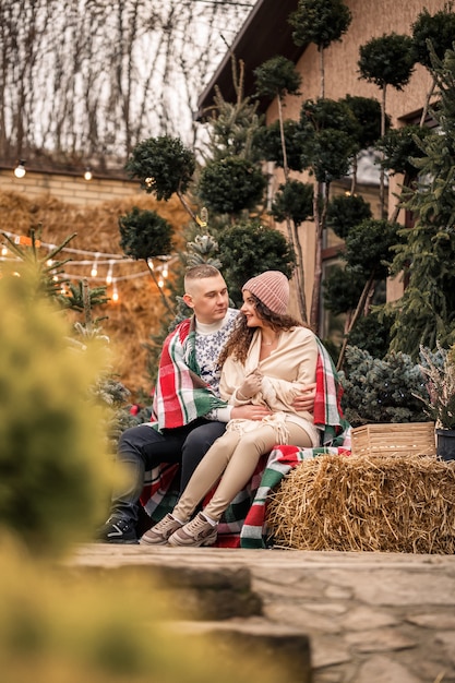 Um lindo jovem casal com roupas brancas está sentado perto de uma árvore de natal no jardim. homem e mulher felizes, romance, celebração de natal, diversão, amor.