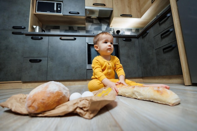 Um lindo menino de 1 ano está sentado na cozinha com pão fresco Criança com pão no chão A criança come pão