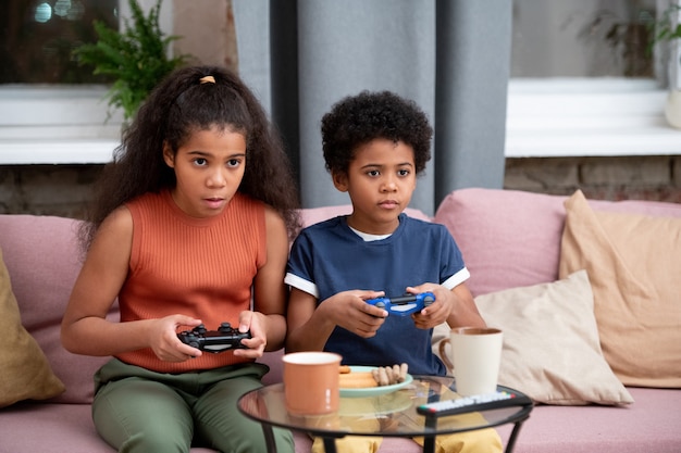 Um lindo menino e uma menina de etnia africana sentados no sofá contra a janela da sala de estar e jogando videogame em frente à TV